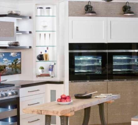 Modern kitchen with a wooden island, built-in oven, Wolf appliance gas stove, shelves with bowls, a wall-mounted TV, and kitchenware on display.