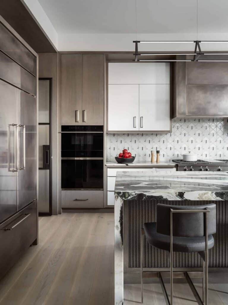 Modern kitchen showroom featuring wood and white cabinetry, marble island, geometric tile backsplash, built-in oven, stainless steel fridge, and a black stool. A luxury Wolf appliance and a bowl of red apples add stylish touches to the space.
