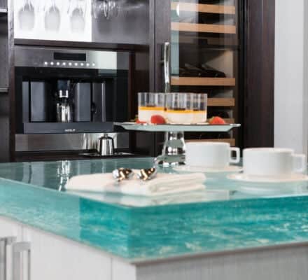 Modern kitchen counter with a thick glass surface, two white coffee cups with saucers, a napkin with utensils, and a cake stand with dessert and fruit—perfectly complemented by a sleek Wolf appliance for that luxury kitchen appeal.