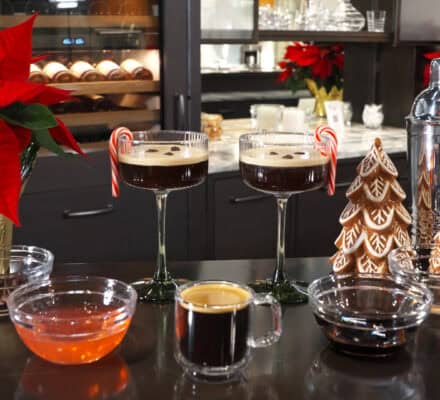 Two glasses of Peppermint Espresso Martini with candy canes, surrounded by ingredients, gingerbread trees, poinsettias, and a cocktail shaker on a kitchen counter.