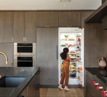 A person stands in a kitchen showroom with dark cabinets and marble countertops, looking inside an open refrigerator filled with food.