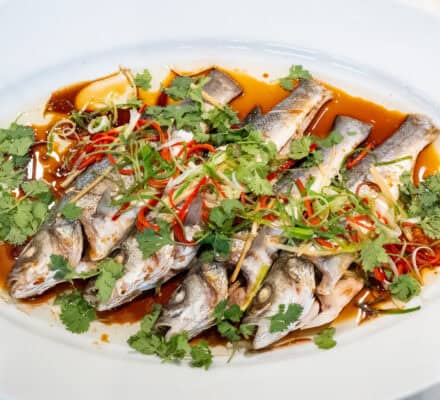 Platter of four whole branzino topped with julienned ginger, scallions, and red Fresno chiles in a savory soy-based sizzling sauce, garnished with fresh cilantro.