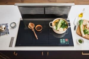 A modern kitchen induction cooktop with a pot of vegetables, wooden spoons, pepper, and herbs; a knife and oil bottle sit nearby on the counter. Discover how to use induction cooktop features for effortless cooking.