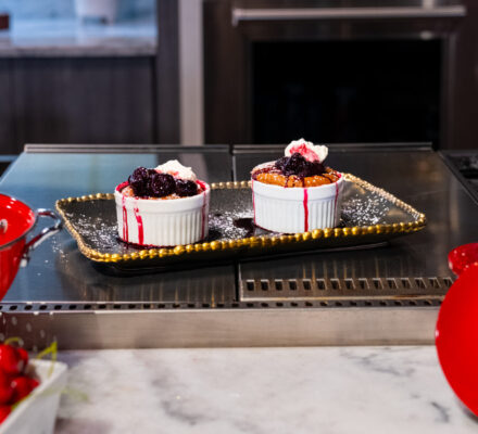 Two berry-topped Dark Chocolate Soufflé with Cherry-Port Reduction in ramekins sit on a tray in a kitchen, with a colander of cherries and a red pot on a marble countertop—perfect paired with cherry-port reduction.