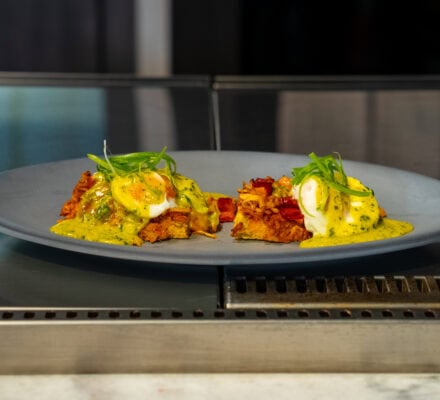 Green Eggs & Hash on a gray plate with two servings of poached eggs on hash browns, topped with yellow sauce and garnished with green onions, makes a delicious breakfast hash on a stainless steel countertop.