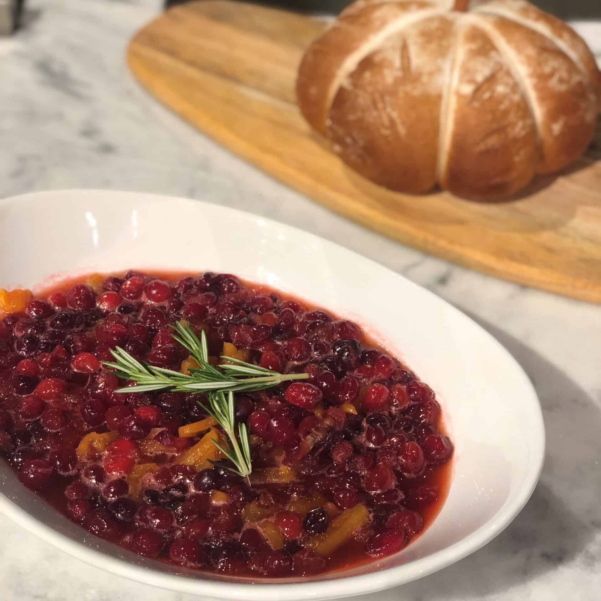 A white bowl of cranberry sauce garnished with rosemary sits on a marble counter, highlighting the elegance of a kitchen featuring Sub Zero appliances, with a round loaf of bread on a wooden board in the background.