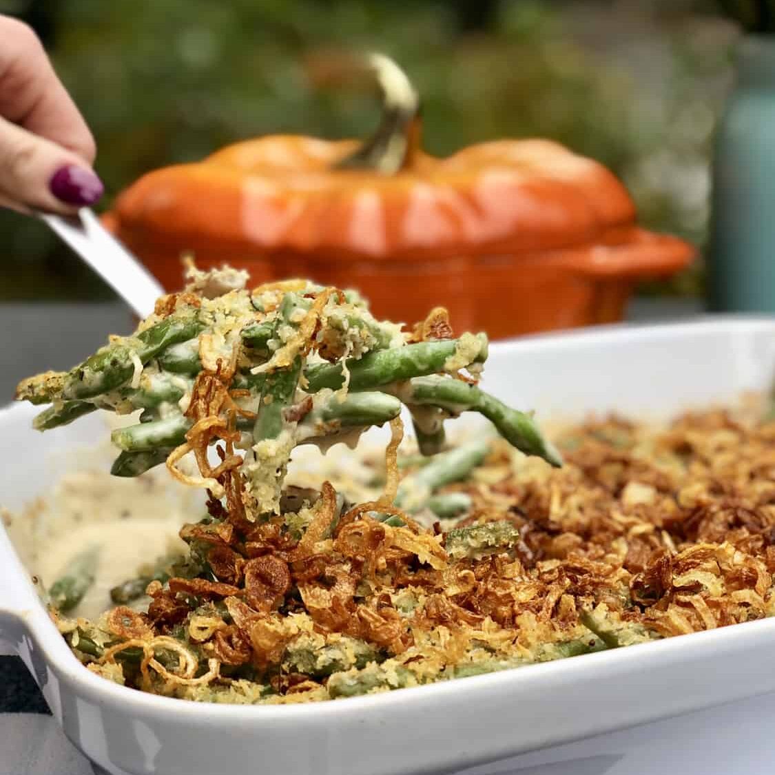 A hand serves green bean casserole topped with crispy fried onions from a white dish in a kitchen showroom; a pumpkin-shaped dish is blurred in the background, highlighting premium Wolf and Sub-Zero appliances.