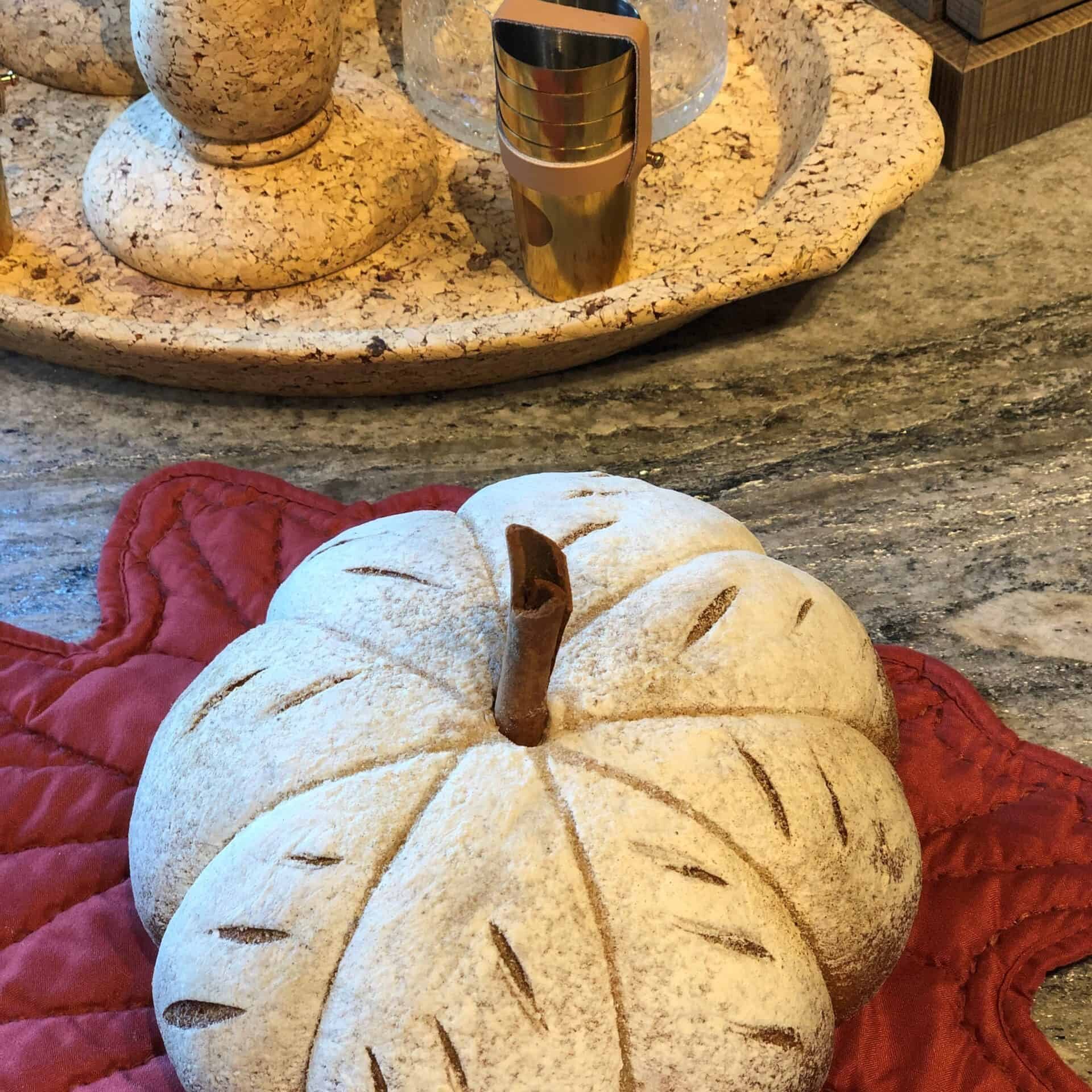 A decorative white pumpkin with brown accents rests on a red leaf-shaped mat atop a granite countertop in a kitchen showroom, with cork coasters and a glass candle holder in the background.