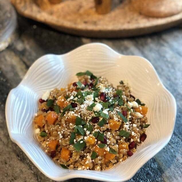A white bowl filled with a grain salad featuring chopped vegetables, herbs, feta cheese crumbles, and pomegranate seeds is placed on a gray countertop in a luxury kitchen appliance showroom.