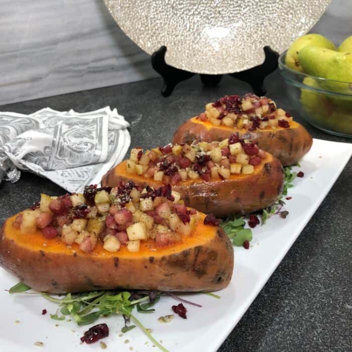 Three halved baked sweet potatoes topped with diced apples, cranberries, and nuts are arranged on a white rectangular plate, styled as they might appear in an elegant appliance showroom display. Pears and a napkin are visible in the background.